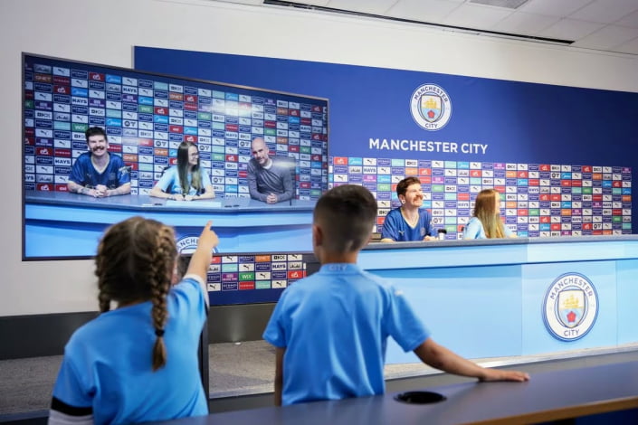 Two children point towards the screen of Pep Guardiola in the press room of the Manchester City stadium during a tour